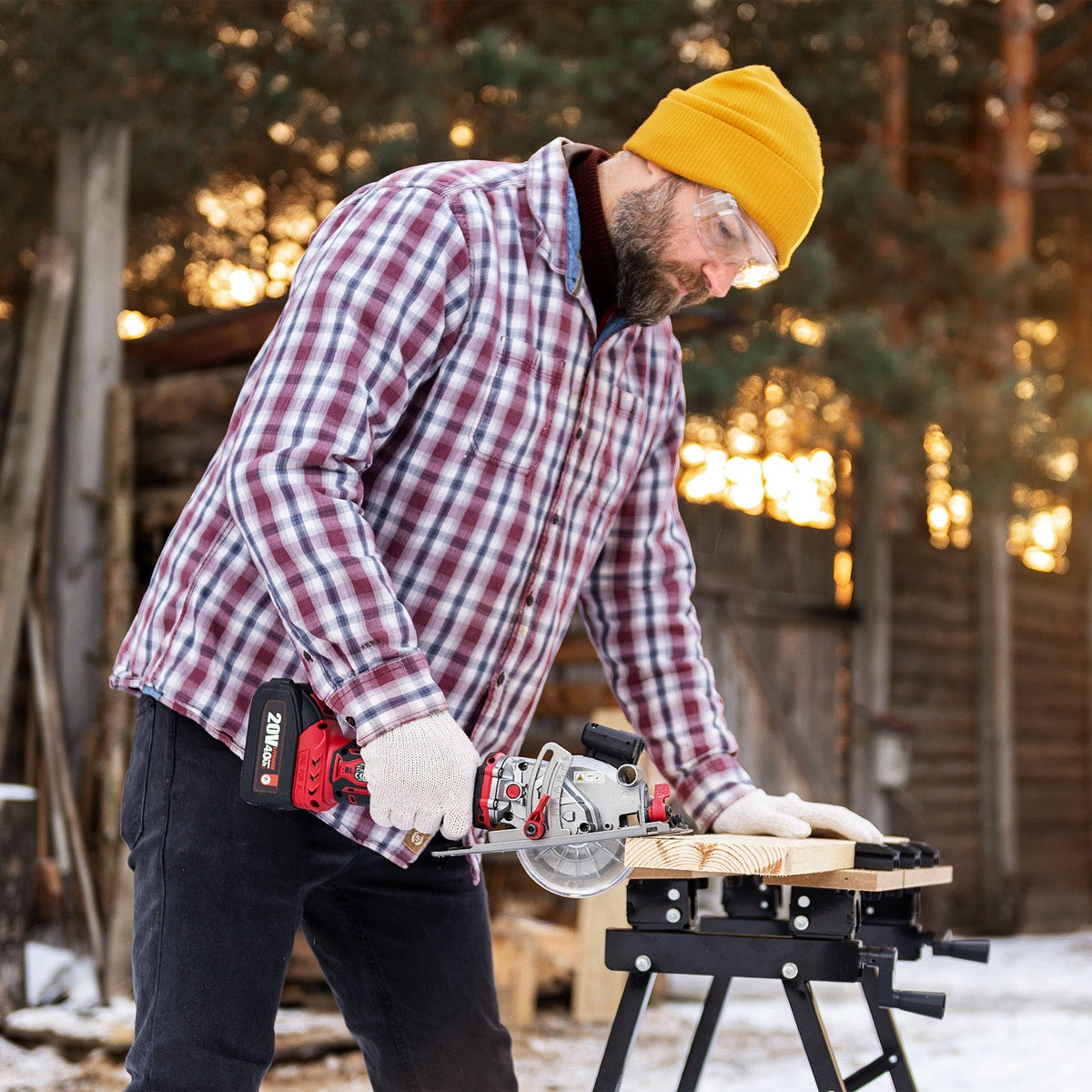 A man uses a Worcraft mini cordless circular saw to cut a piece of wood on a black sawhorse outdoors in a snowy backyard.