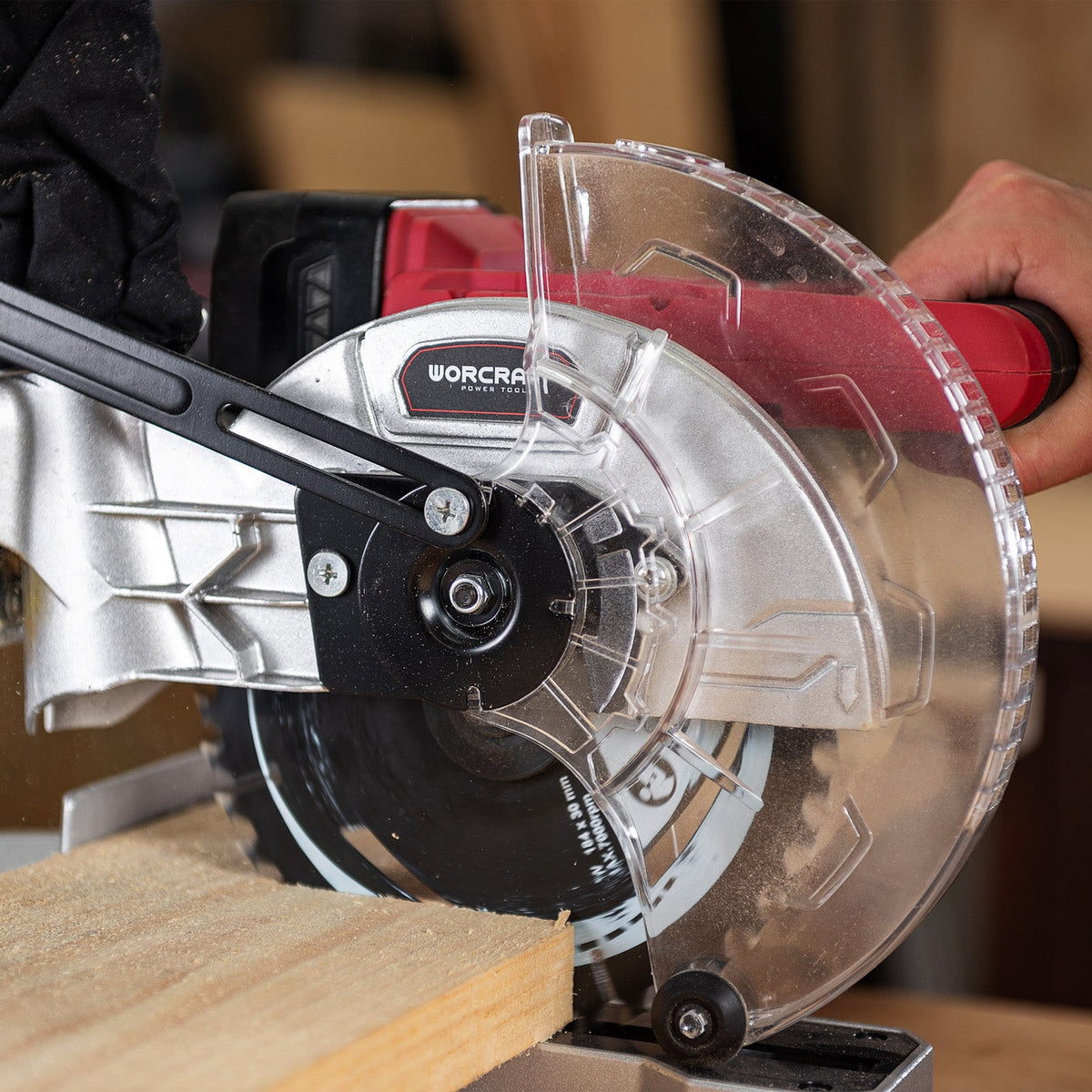 A close-up of a red and black Worcraft circular saw with a clear protective guard, cutting a piece of wood. A person's hand is visible holding the saw handle.