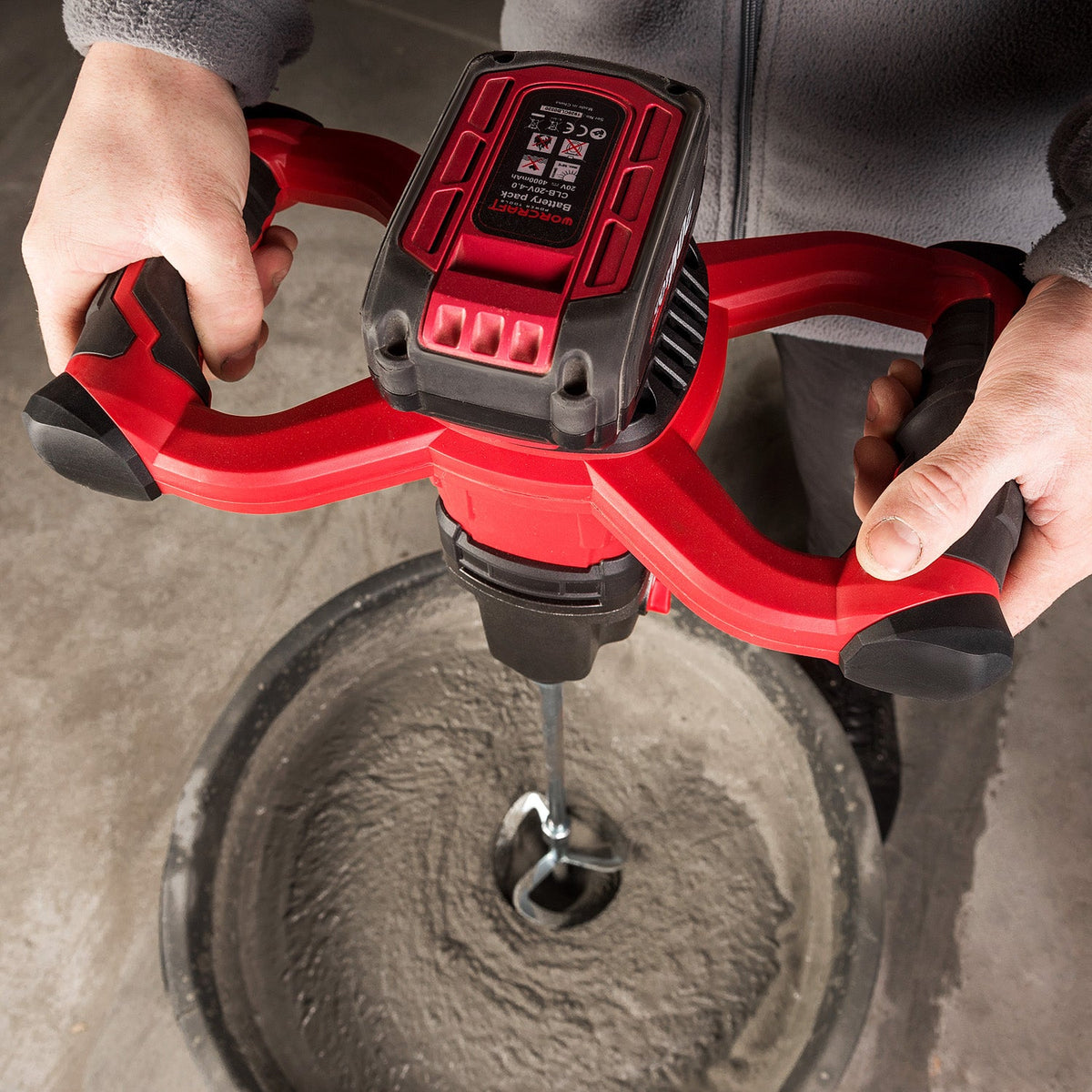 A cordless Worcraft wet mix drill being used by a person to mix concrete in a bucket. The drill has a red and black handle with a digital display on the battery pack, and the mixing paddle is submerged in wet gray concrete.