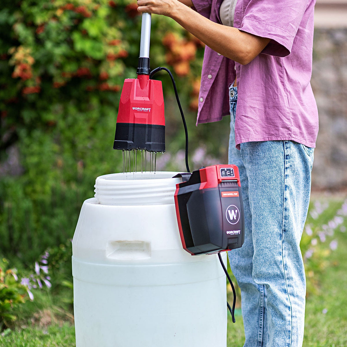 A person outdoors uses a red and black Worcraft cordless attachment with a long metal wand to mix contents in a large white plasticrain barrel.