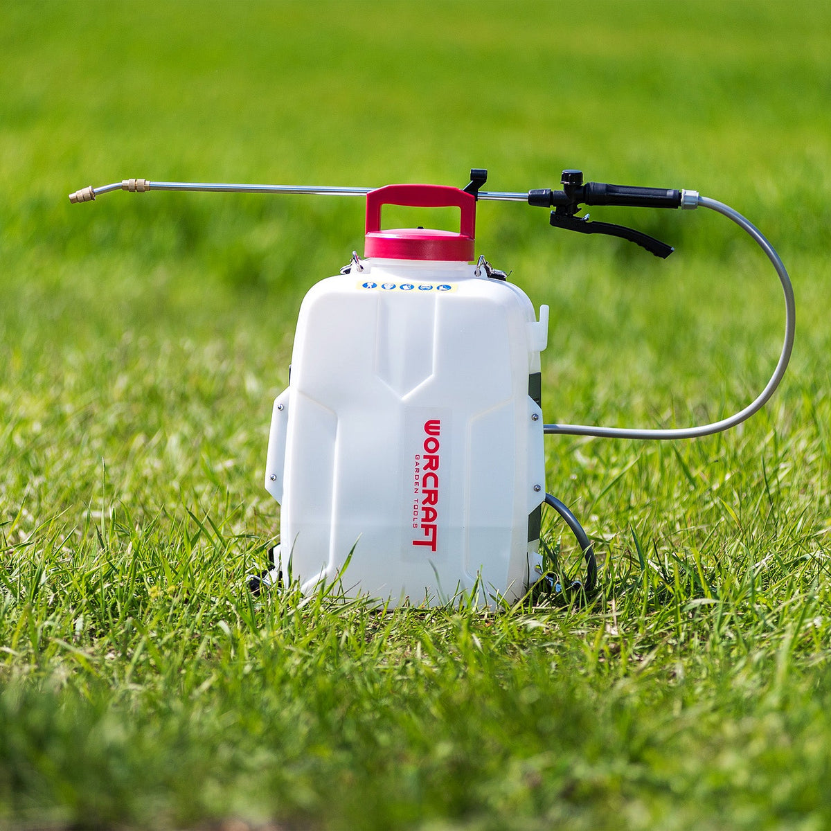 A white Worcraft backpack sprayer with a red cap and a long metal spray wand, set on a green grass lawn.