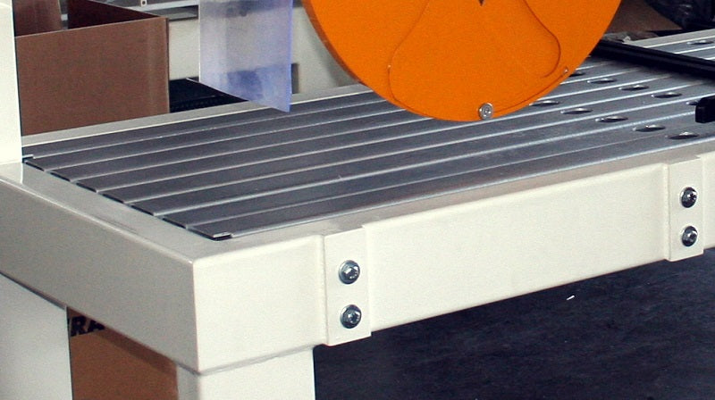 Close-up of a metal slatted worktable or conveyor surface with a white frame, an orange circular component above, and a dark floor underneath.